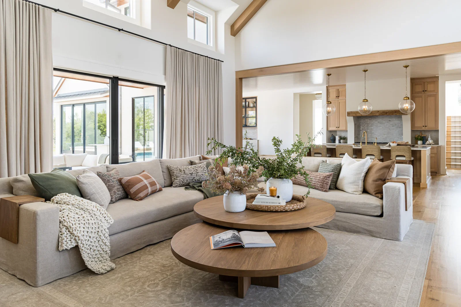 Living room with light-colored sofa, multiple pillows, wooden coffee tables with decorative vases and a large open kitchen in the background with pendant lights, wooden cabinets, and a marble fireplace.