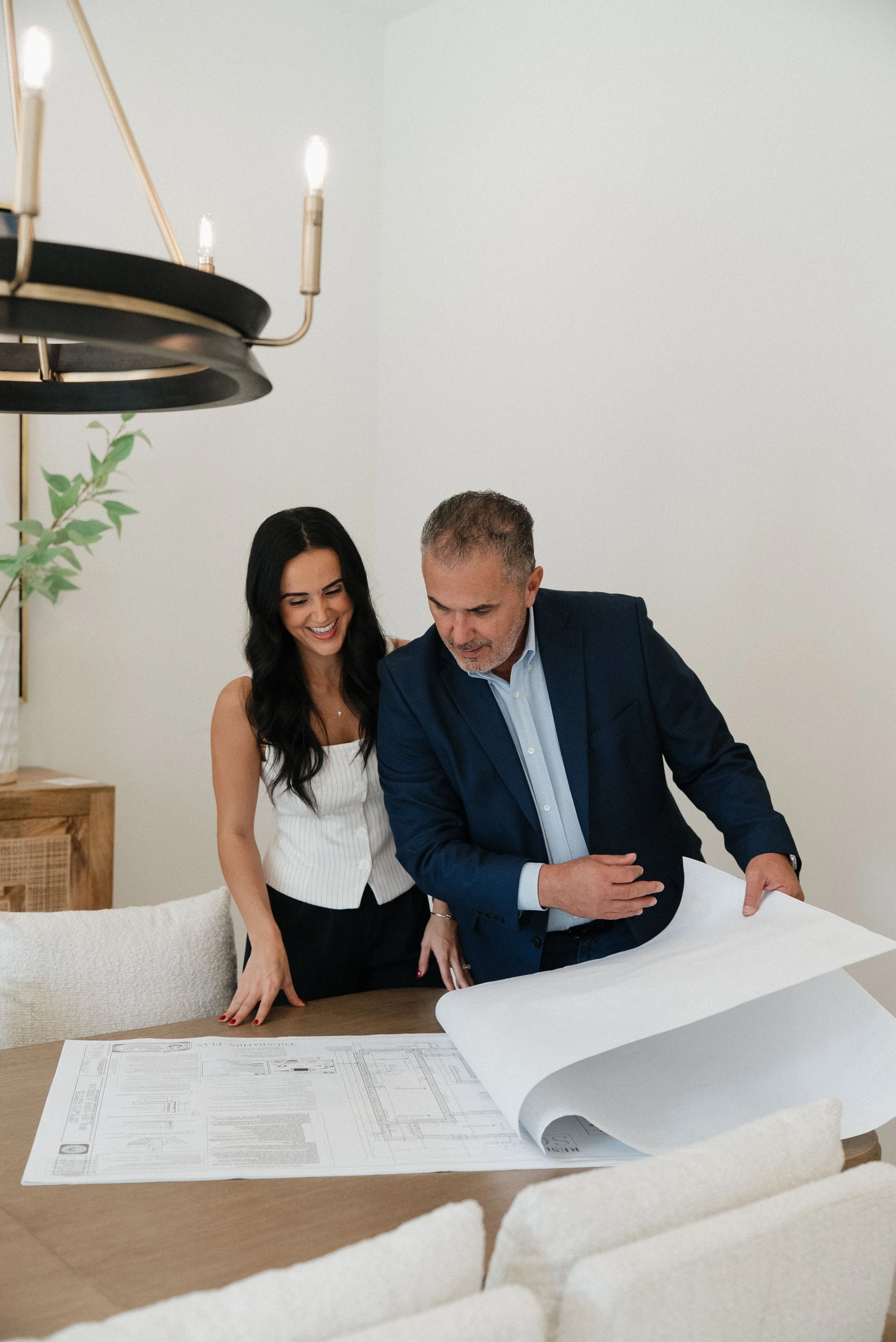 Two people, a woman and a man, are looking at architectural blueprints spread out on a table. They seem to be discussing the plans in a well-lit room with a modern chandelier and a green plant in the background.