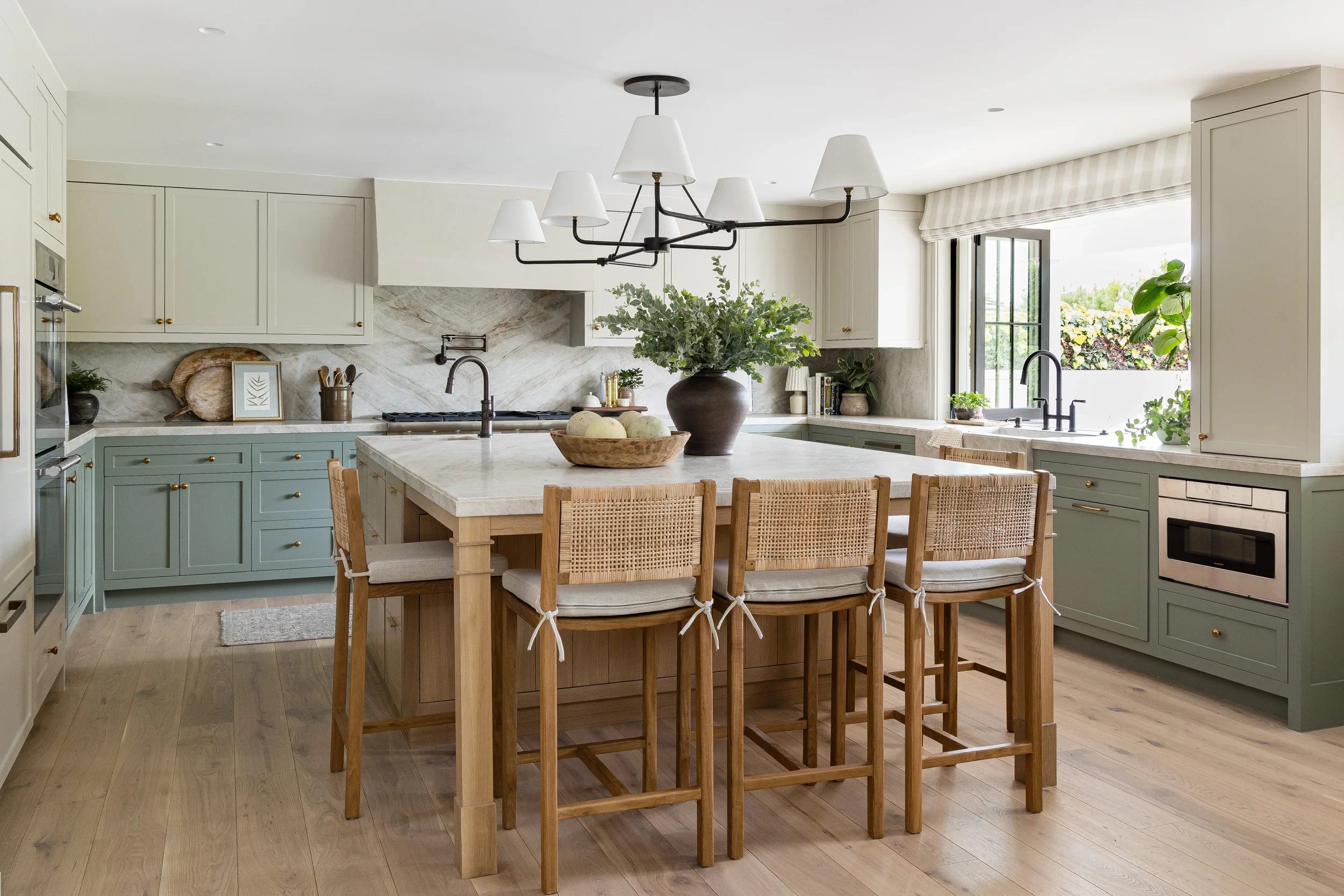 A modern kitchen with a central island, light green and white cabinetry, a marble backsplash, and a black chandelier with white lampshades hanging above the island. There are plants and books near the window, which opens to a garden scene.