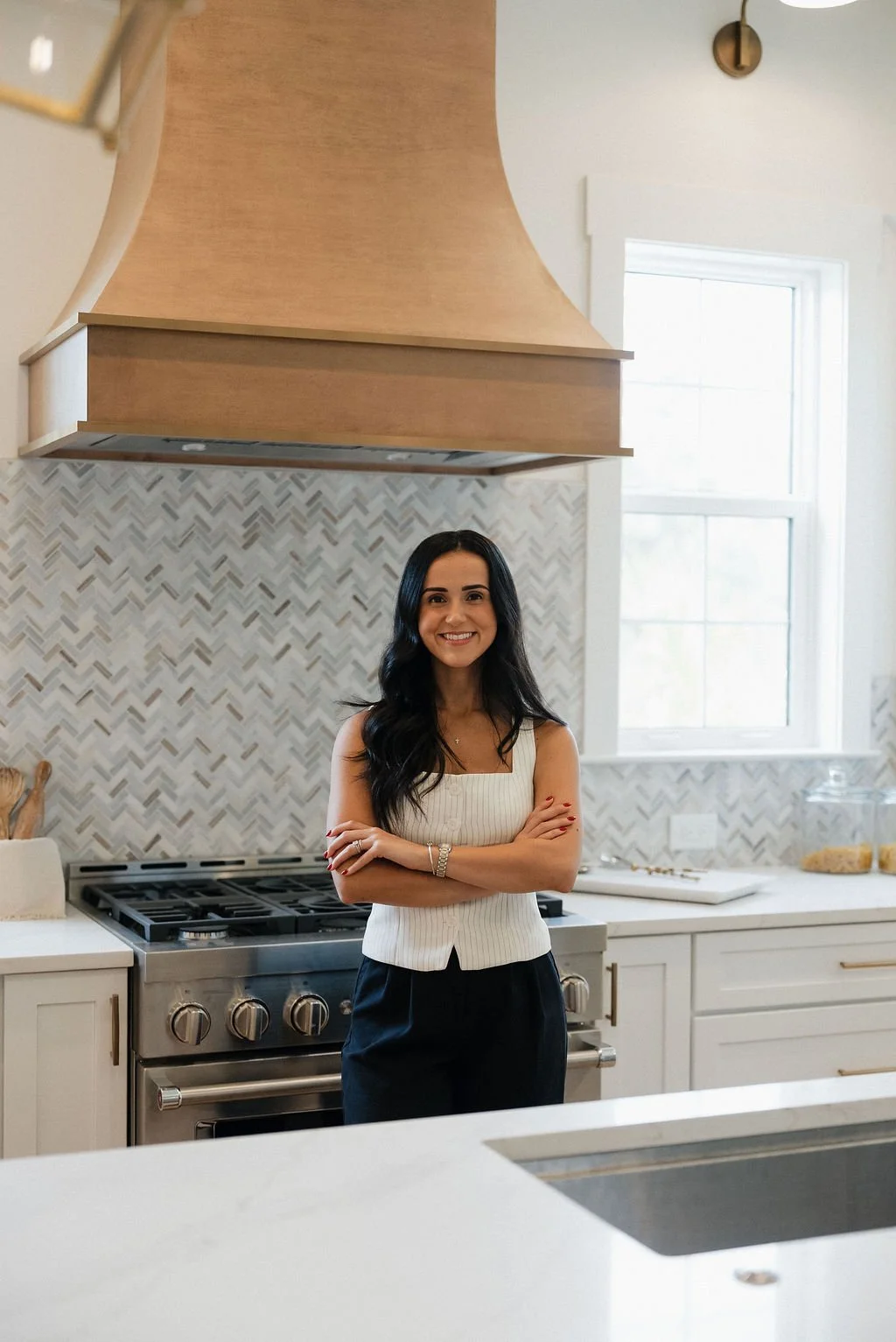 A woman with long dark hair smiling and crossing arms, standing in a modern kitchen with white cabinetry, a stainless steel stove, and a light-colored patterned tile backsplash.