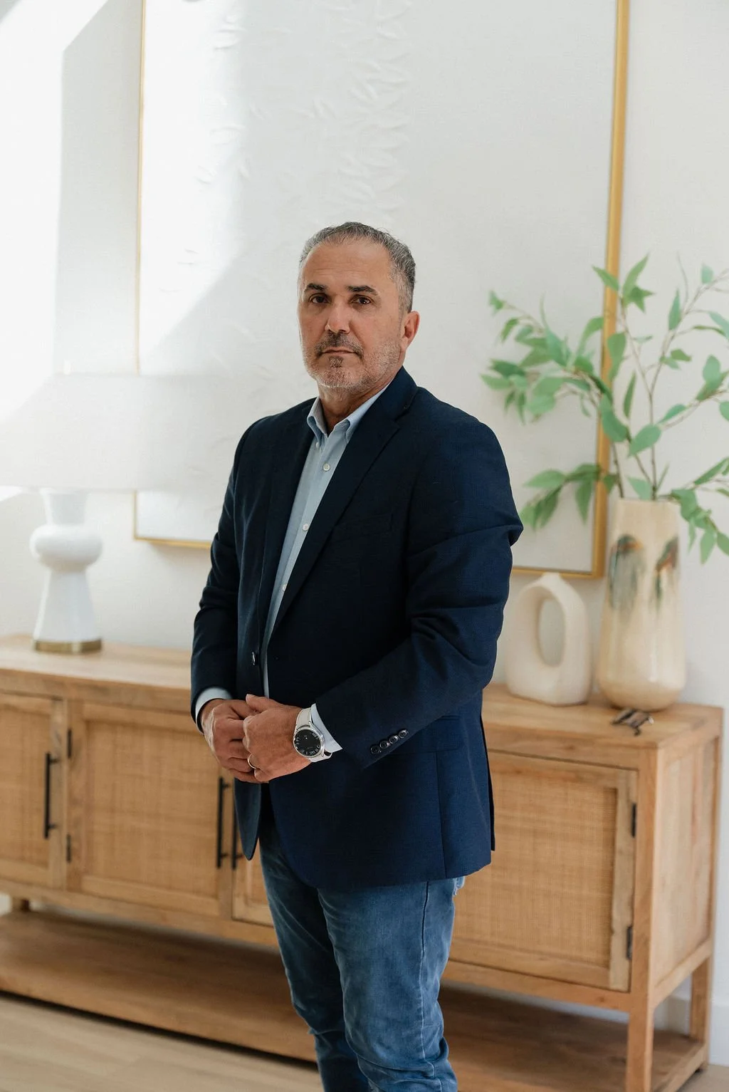 A middle-aged man with gray hair and a beard wearing a navy blazer, light blue shirt, and jeans, standing indoors in front of a wooden cabinet, decorative vases, and a large potted plant.