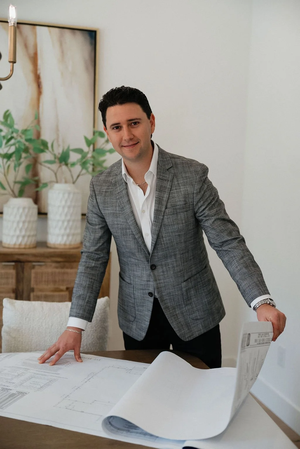 A man in a gray plaid blazer and white shirt standing at a table with blueprints, in a modern room with potted plants and artwork on the wall.