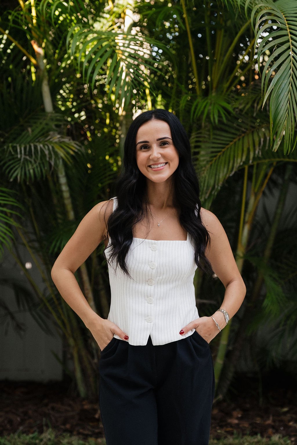 A woman with long black hair, wearing a white sleeveless top with vertical stripes and black pants, stands outdoors in front of lush green palm trees, smiling at the camera.