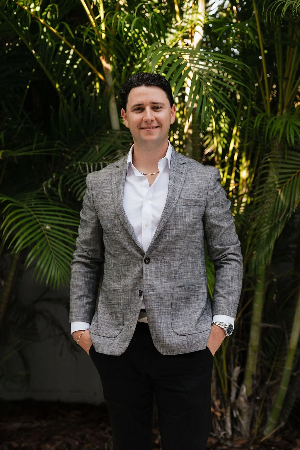 A young man in a gray plaid blazer and white shirt standing outdoors in front of lush green tropical plants, smiling with hands in his pockets.