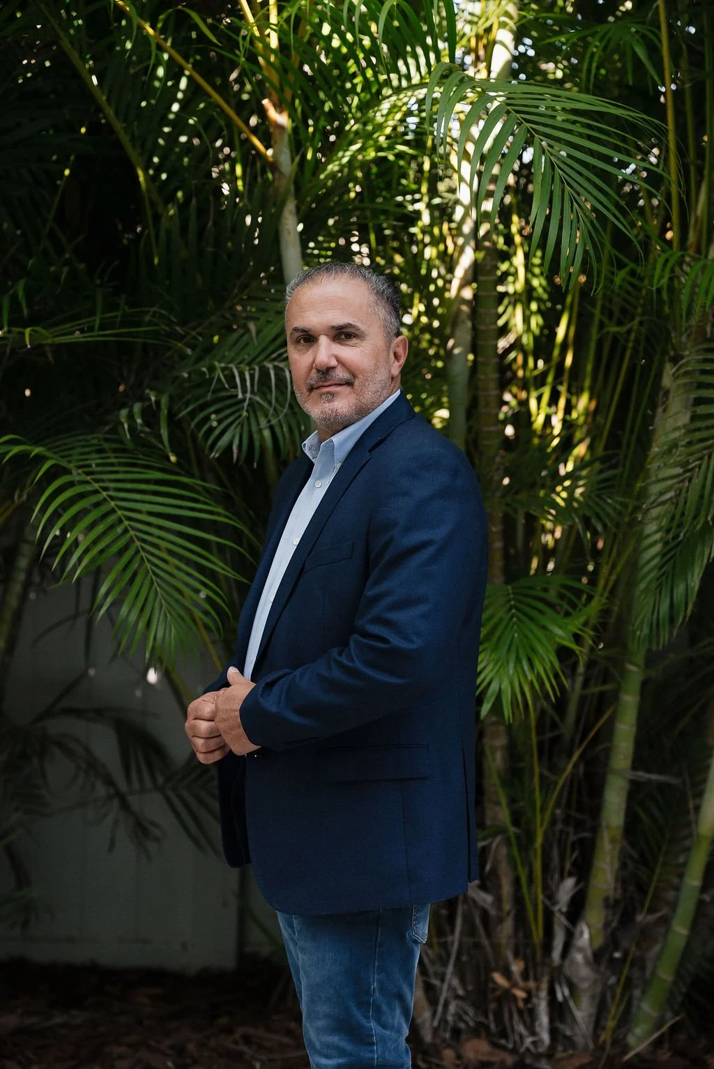 A middle-aged man in a navy blazer and light blue shirt stands outdoors in front of lush green tropical plants, looking at the camera.