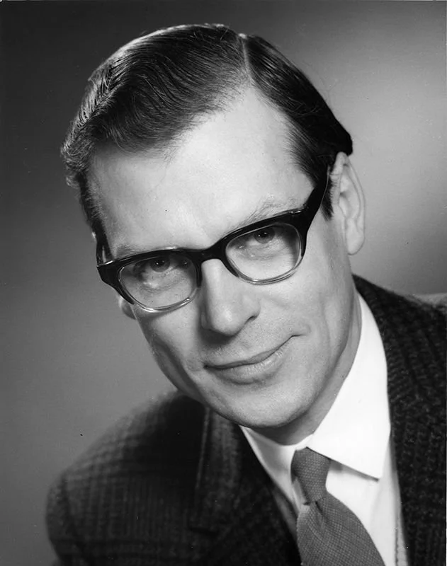 Black and white portrait of a man wearing glasses, a suit, and a tie, with neatly combed hair, smiling slightly.