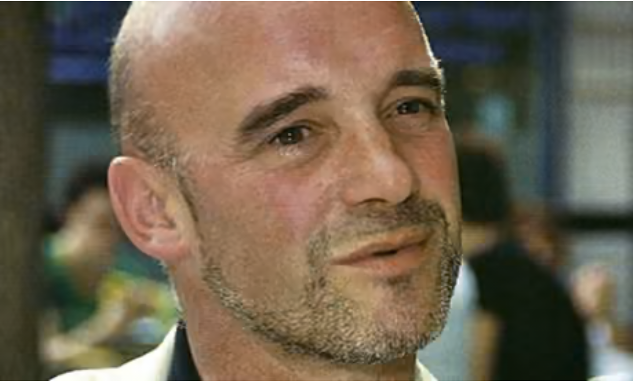 Close-up of a bald man with a short beard, smiling softly, in a crowded indoor setting.