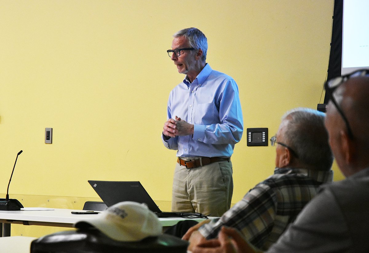 A middle-aged man with glasses, wearing a light blue dress shirt and beige pants, is standing and speaking during a presentation. Several people are seated in front of him, listening, with a yellow wall background and a partially visible screen to the right.