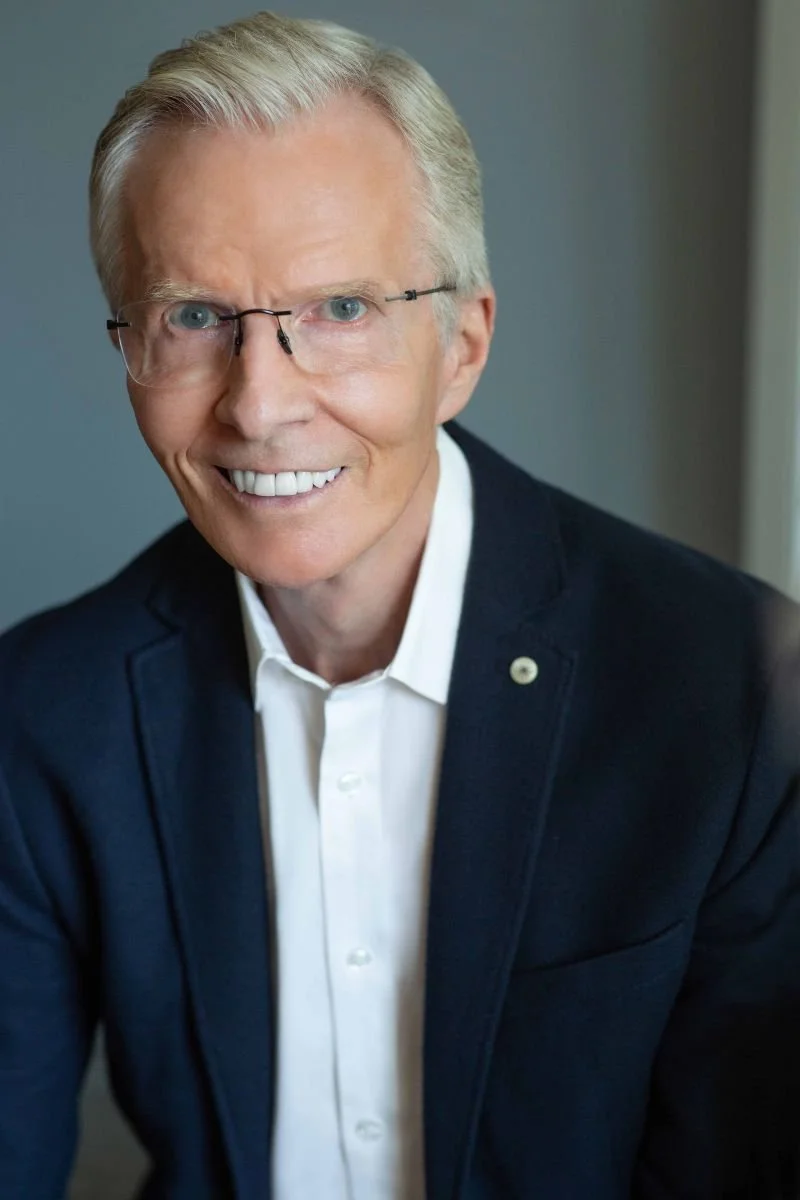 A smiling older man with blond hair, wearing glasses, a white shirt, and a dark blazer, in a professional setting.