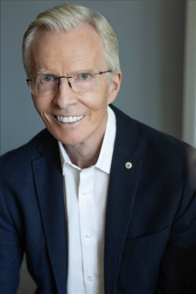 A smiling older man with blonde hair, wearing glasses, a white shirt, and a dark blazer, sitting indoors against a gray background.