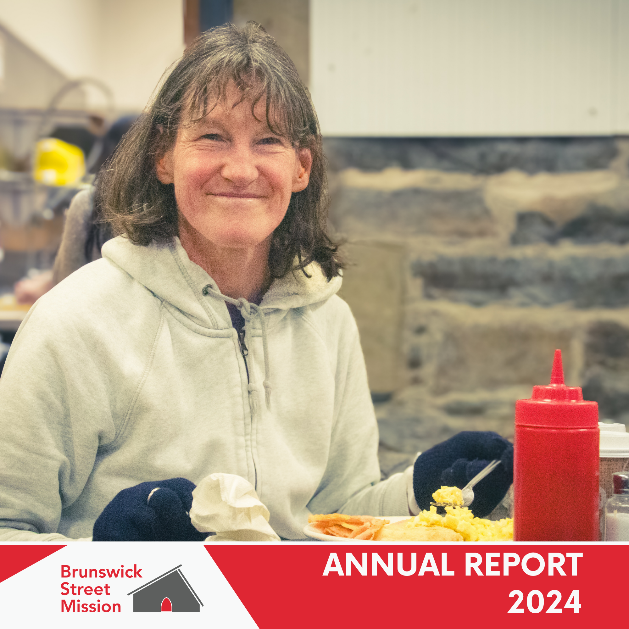 A woman with shoulder-length brown hair, smiling, sitting at a table with breakfast food, a plate with eggs, ham, and toast, ketchup bottle, and coffee, in a rustic setting. She is wearing a light gray hoodie and black gloves.