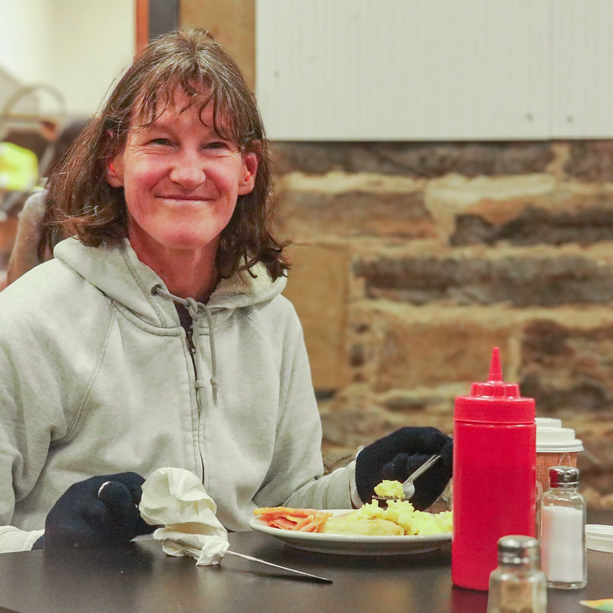 A woman with brown wavy hair wearing a light-colored hoodie, sitting at a table with what appears to be mashed potatoes, slices of ham, and other food items, near bottles of condiments including ketchup, in a room with stone wall decor.