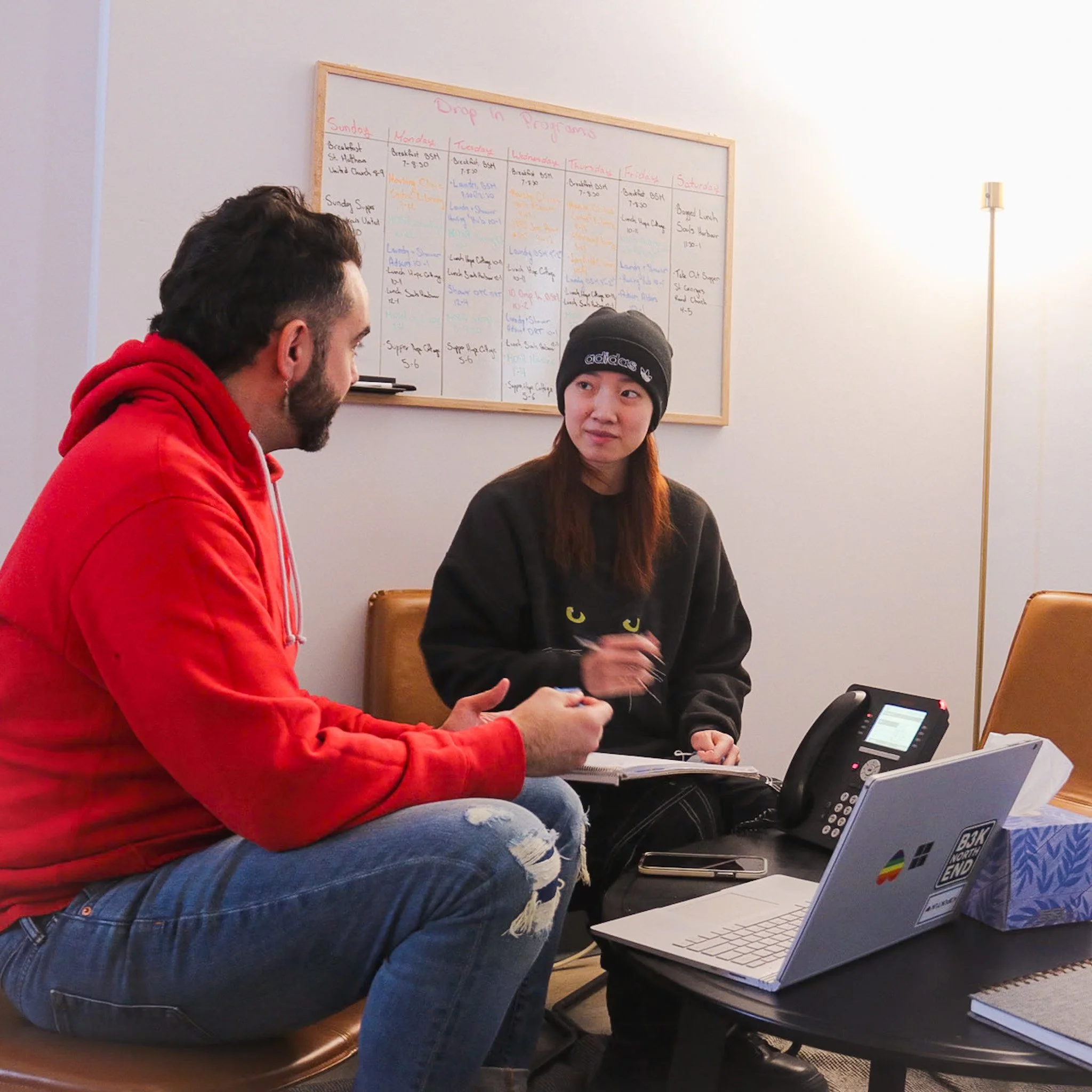Two people sitting and talking in front of a whiteboard with a weekly schedule, with laptops, a phone, and a notebook on the table.