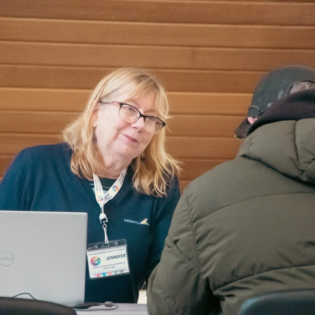 A woman with blonde hair, glasses, and a lanyard sitting at a table, speaking with a person wearing a helmet and a green jacket, against a wooden wall background.