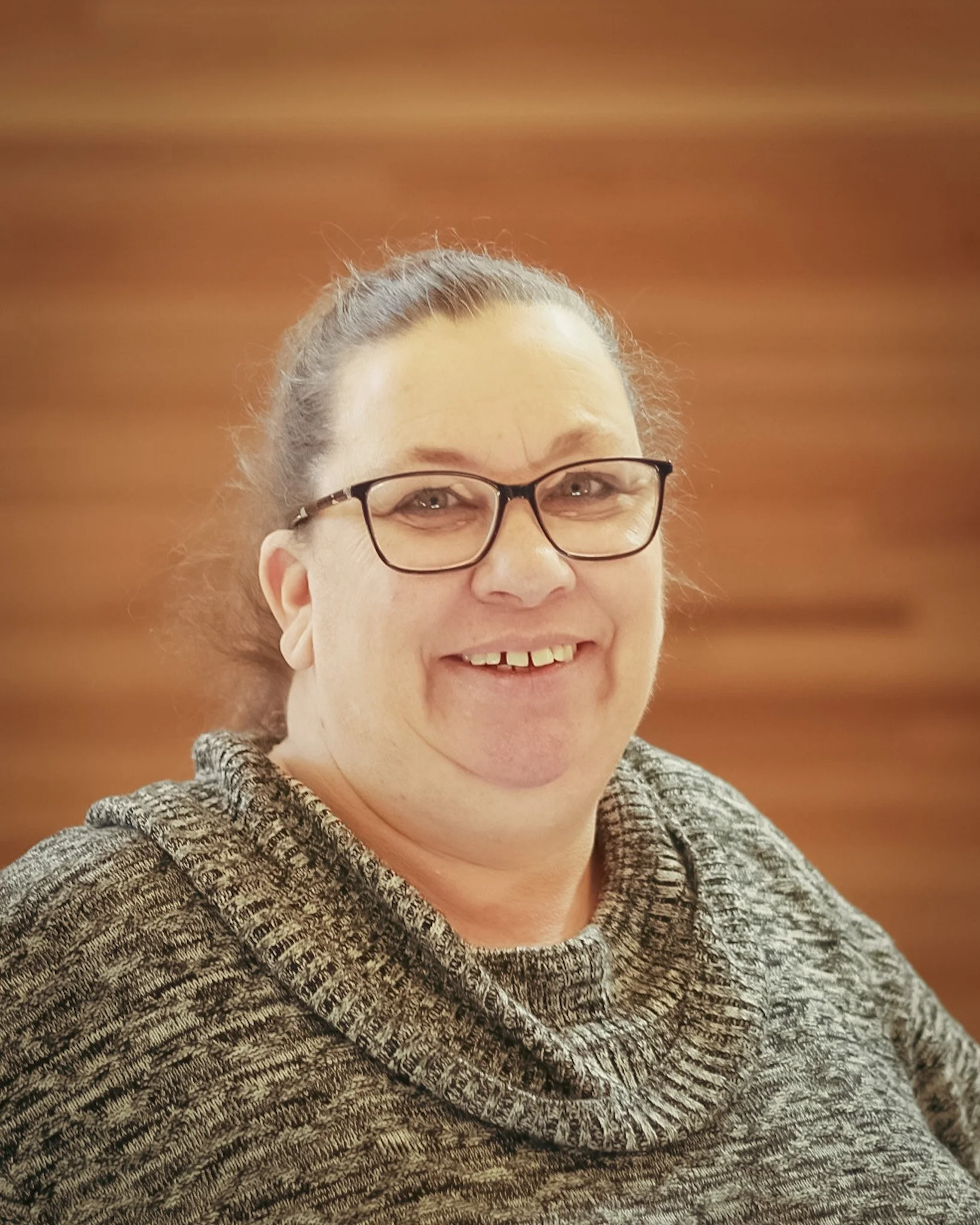 A woman with glasses and brown hair tied back, smiling and wearing a marled gray sweater, against a wooden background.