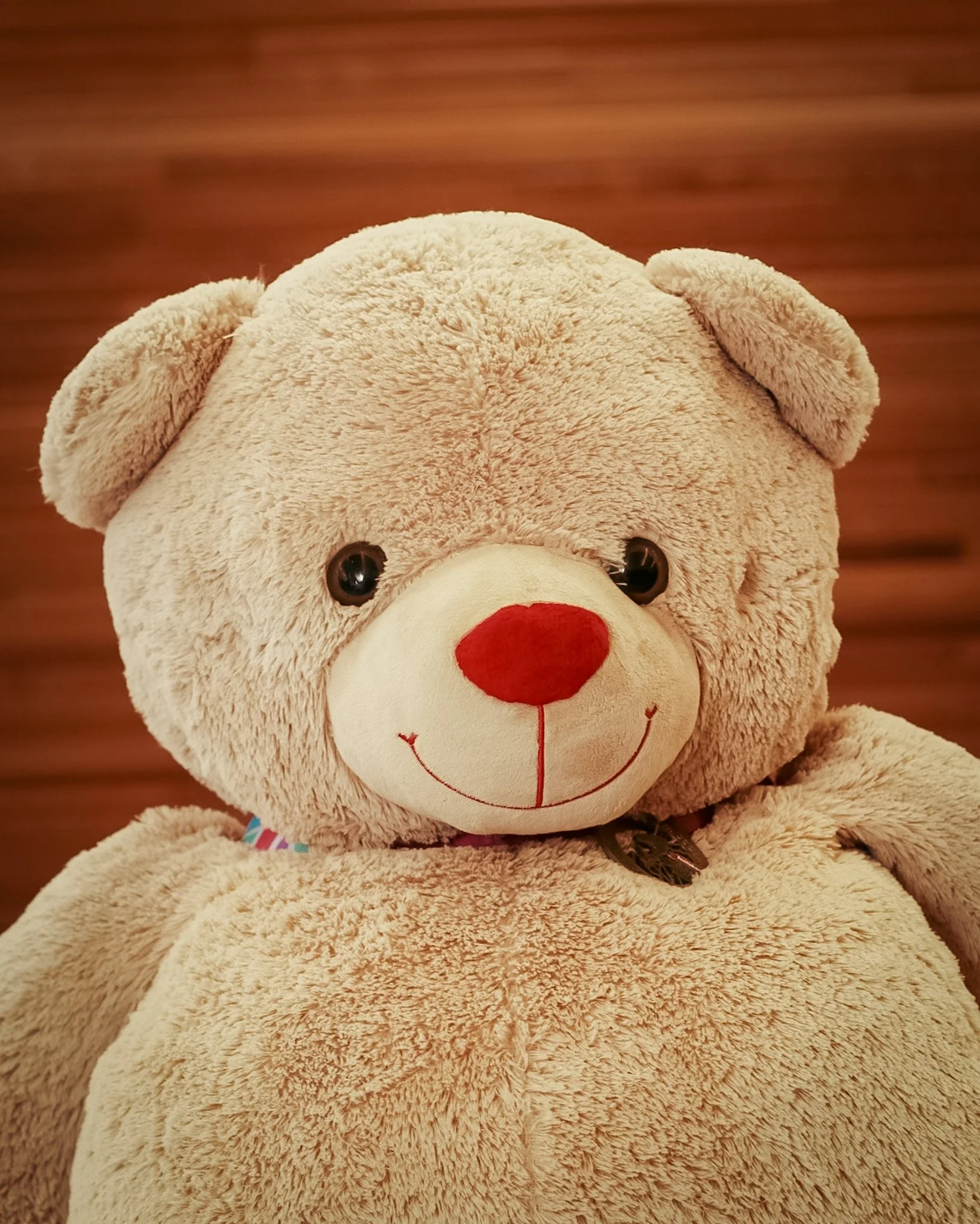 A close-up of a light brown teddy bear with black eyes, a red nose, and a smiling face, sitting against a wooden background.
