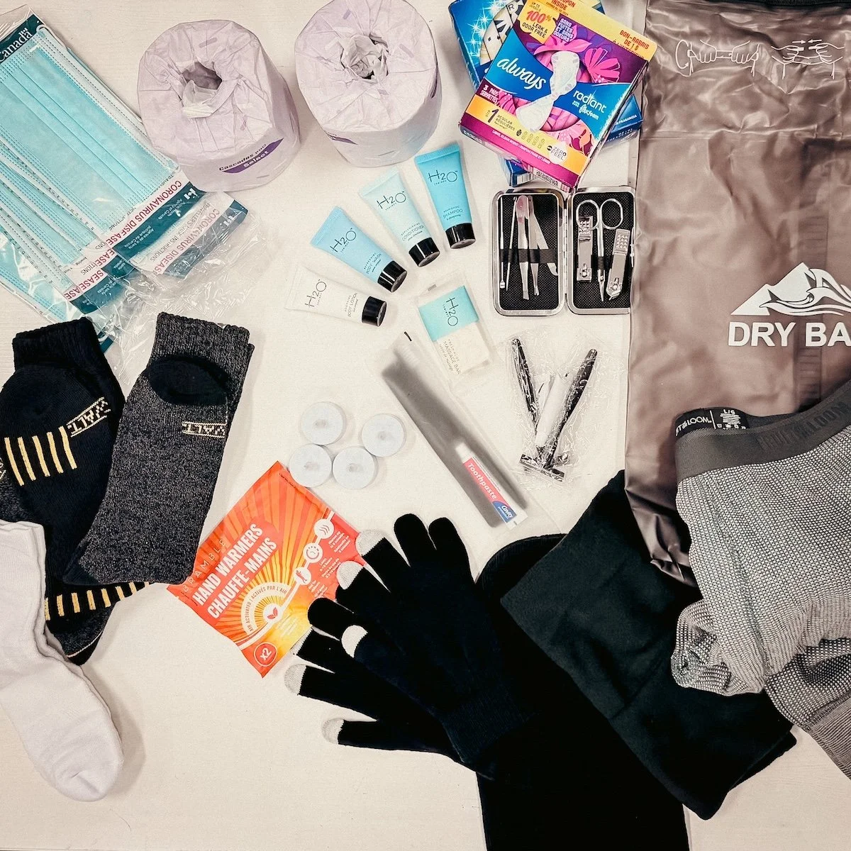 First aid supplies and personal protective equipment, including face masks, gloves, antiseptic wipes, and medical tools, are laid out on a white surface. There are also toiletries such as toothpaste and tissues, and a brown dry bag for outdoor activities.