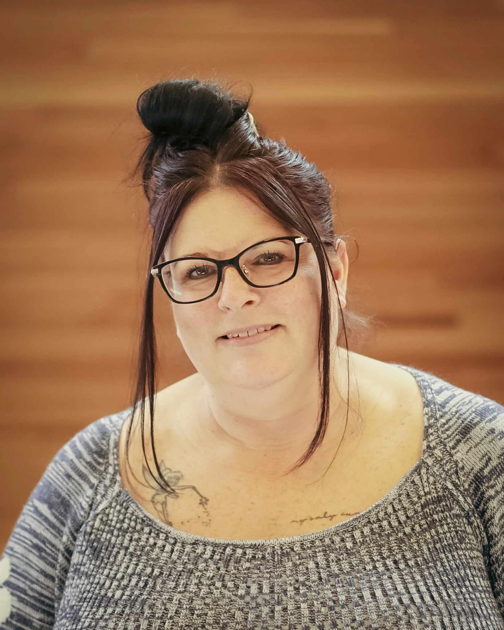 A woman with dark hair in a top knot, glasses, and a tattoo on her shoulder, smiling at the camera with a wooden background.
