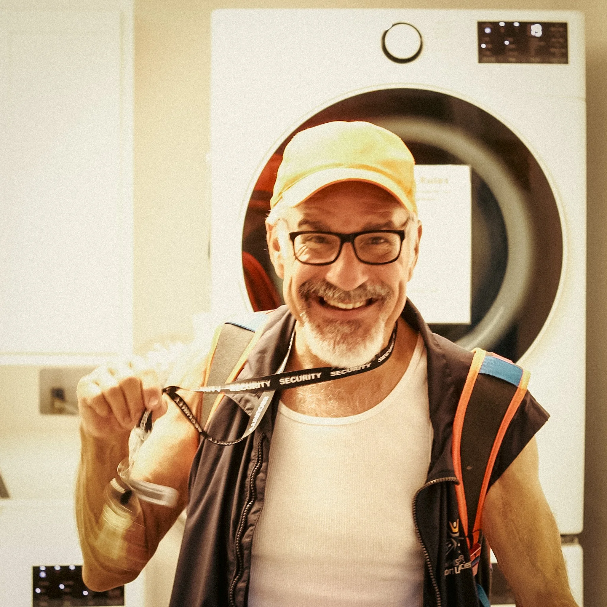 A man with glasses wearing a baseball cap and a backpack, smiling as he stands in front of a washing machine.