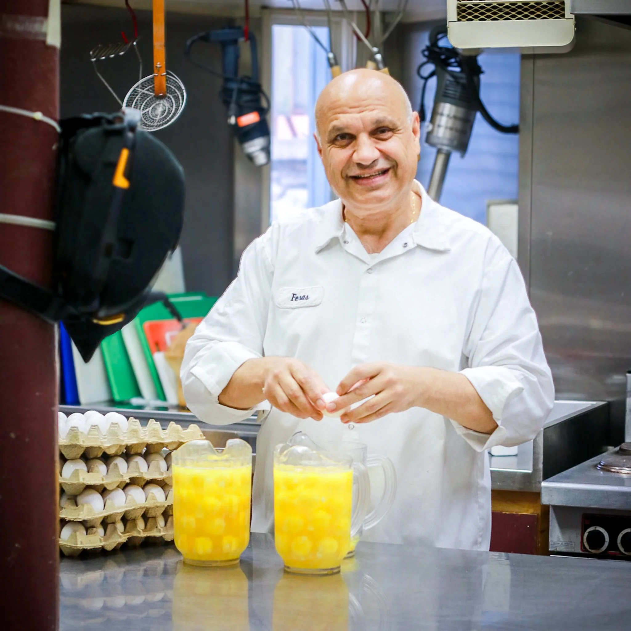 A man with a shaved head and white chef coat standing in a commercial kitchen, smiling at the camera. He is surrounded by kitchen equipment and ingredients, including three glasses of yellow liquid on the counter in front of him.