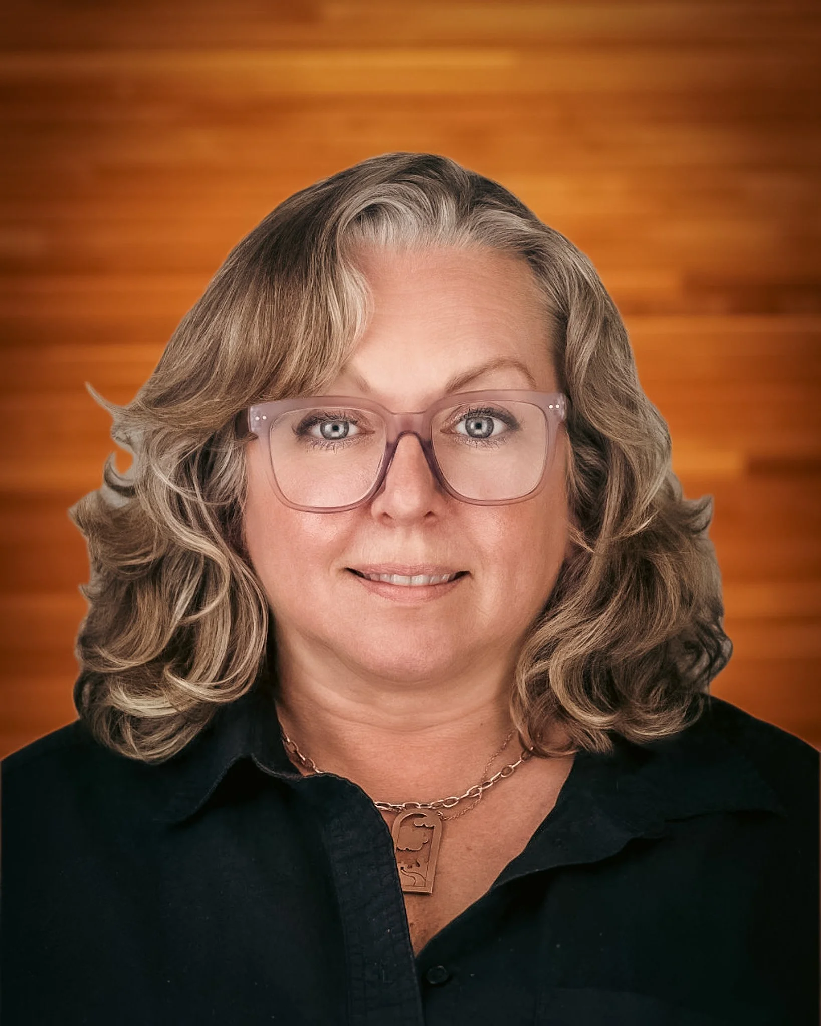 A woman with wavy blonde hair, wearing large clear glasses, a black shirt, and a gold necklace with a dog pendant, smiling against a wooden background.