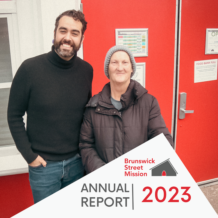 A man and woman standing together, smiling, with a red wall and bulletin boards behind them, holding an annual report for Brunswick Street Mission 2023.
