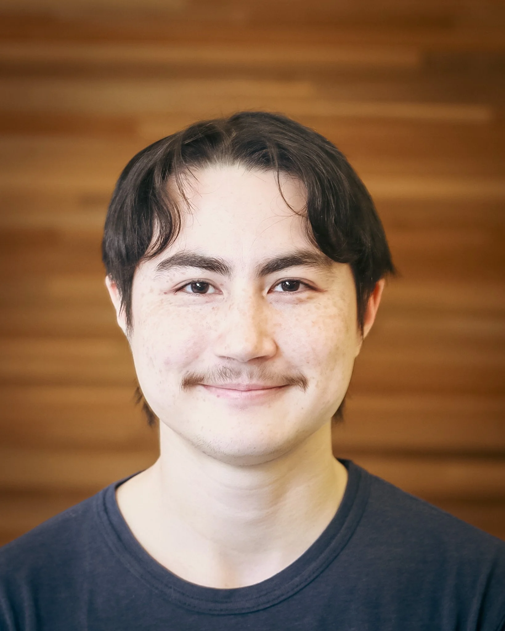 Close-up of a smiling young person with short dark hair, freckles, and light skin, wearing a black T-shirt, against a wooden background.