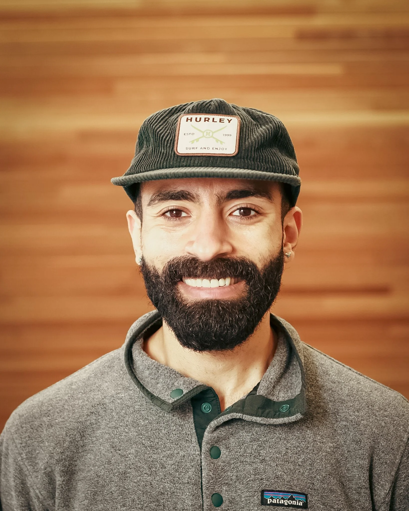 A man with a dark beard and mustache, wearing a gray Patagonia shirt and a dark green hurley cap, smiling in front of a wooden wall background.