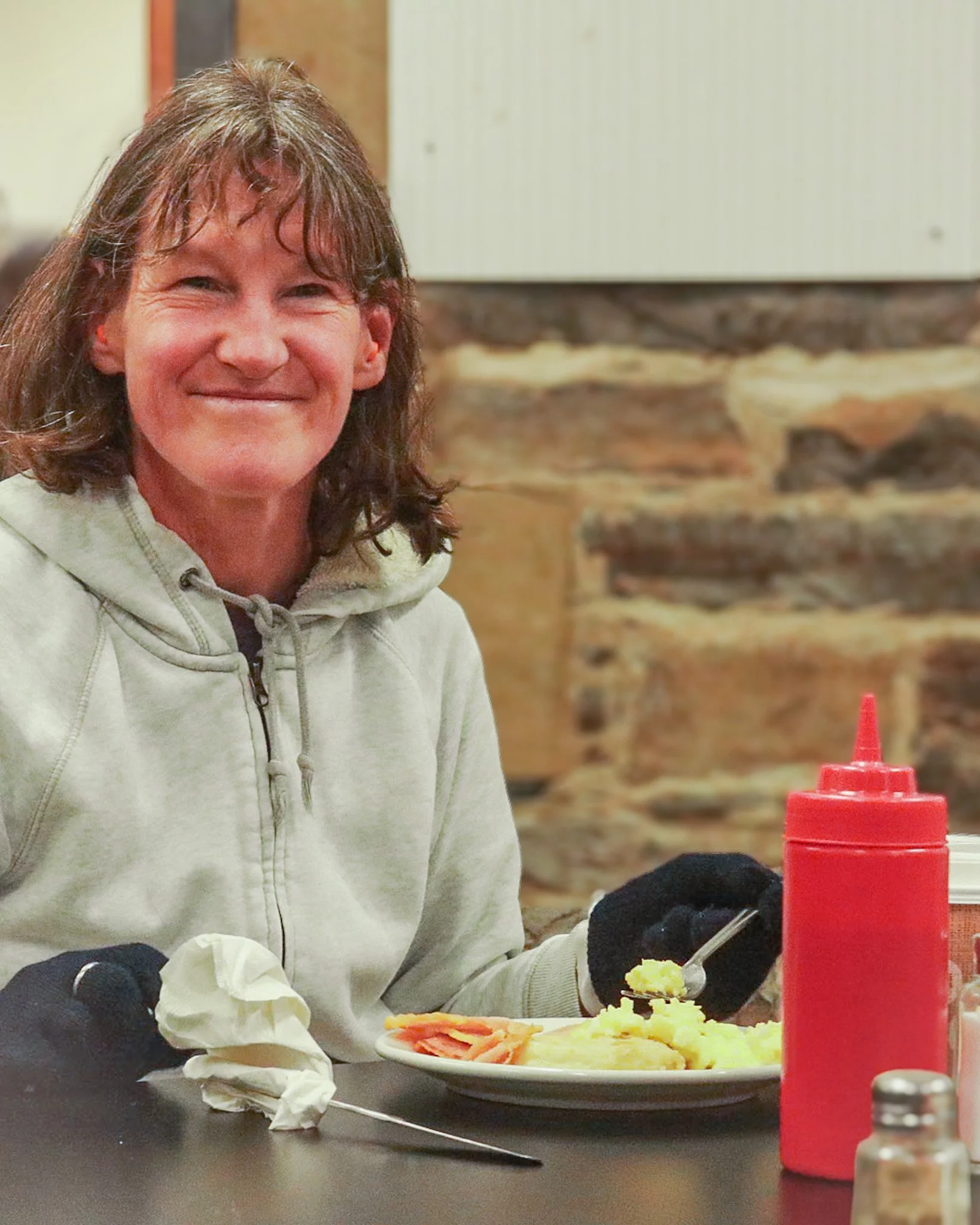 Woman smiling in a casual restaurant, wearing a light-colored hoodie, sitting at a table with food, ketchup bottle, and tissue.