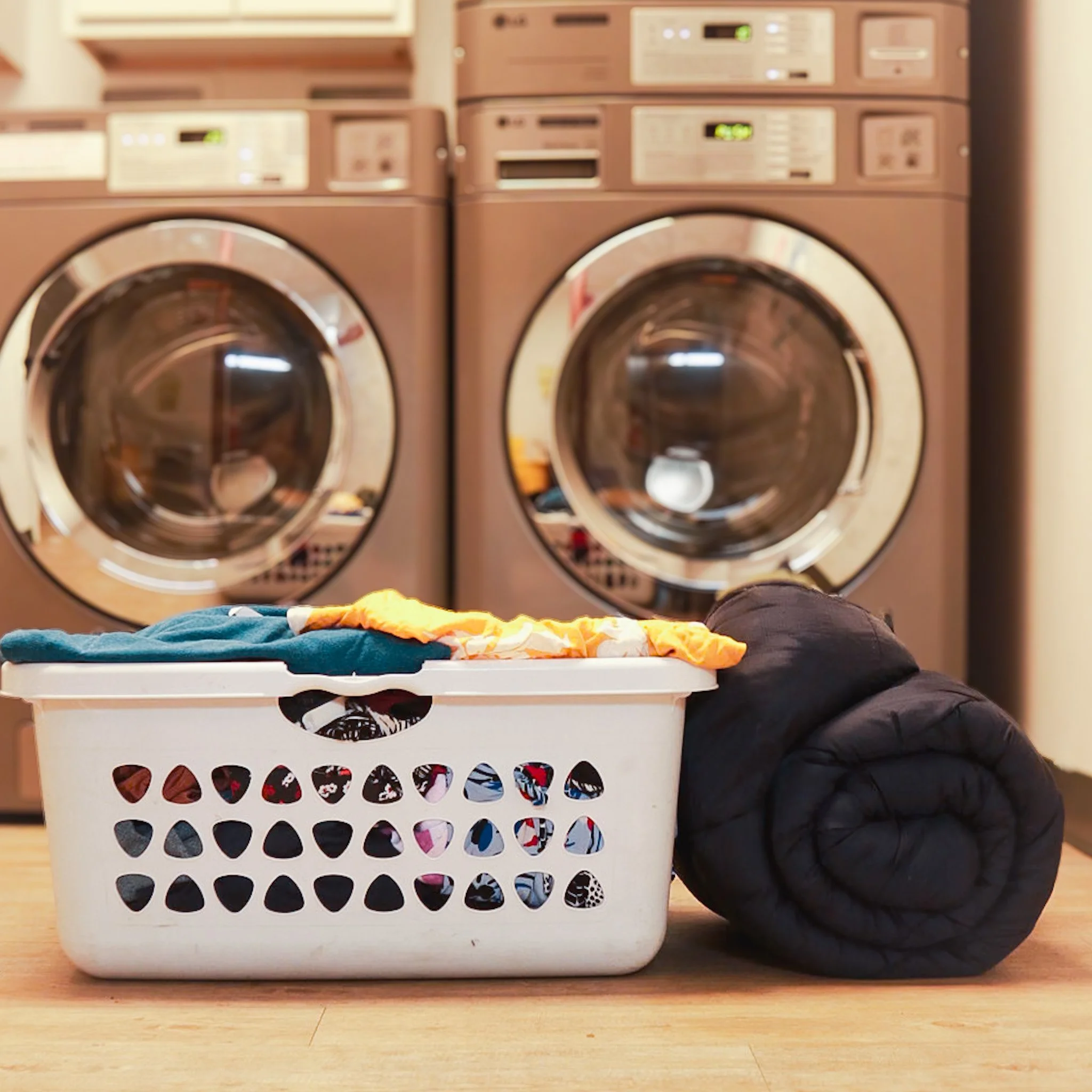 A laundry basket filled with clothing, placed on the floor in front of two stacked front-loading washing machines.