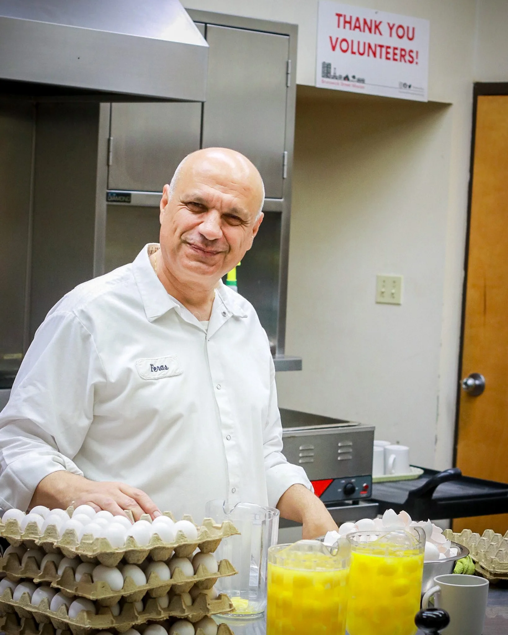A man in a white shirt with a name tag 'Feras' faces the camera, standing behind a table filled with cartons of eggs and yellow juice glasses, in a kitchen setting. A sign on the wall reads 'Thank you volunteers!'