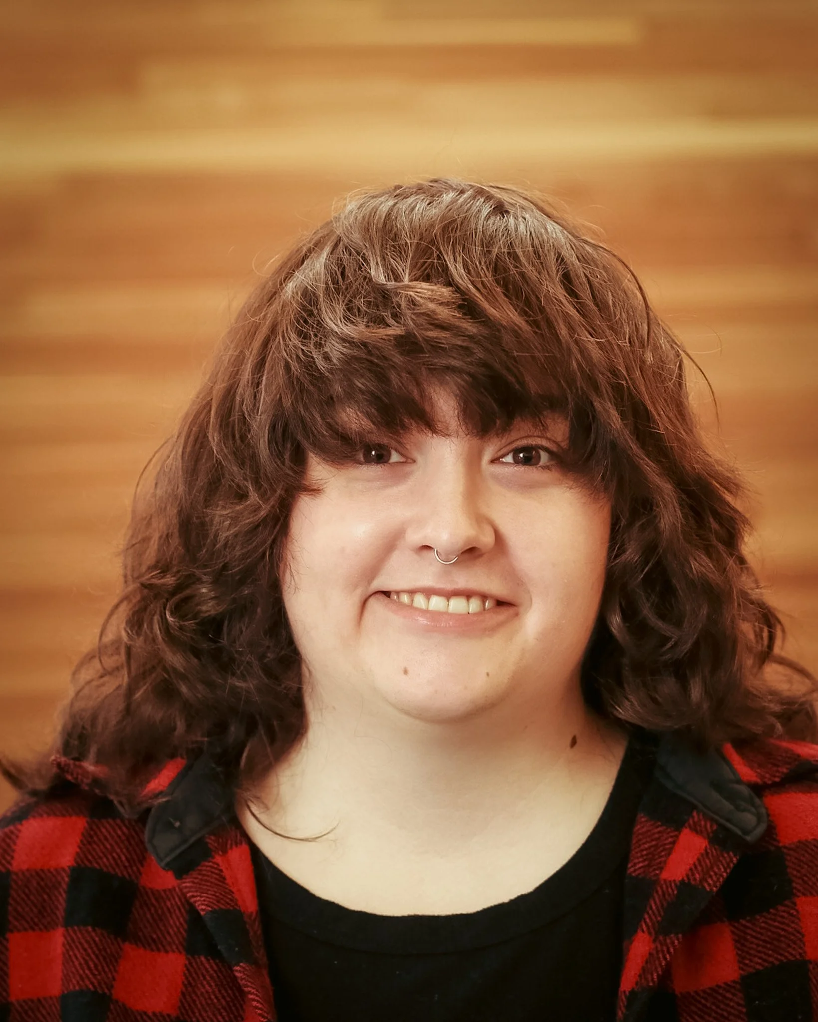 Young man with wavy brown hair, smiling, wearing a red and black checkered shirt and sitting in front of a wooden wall background.