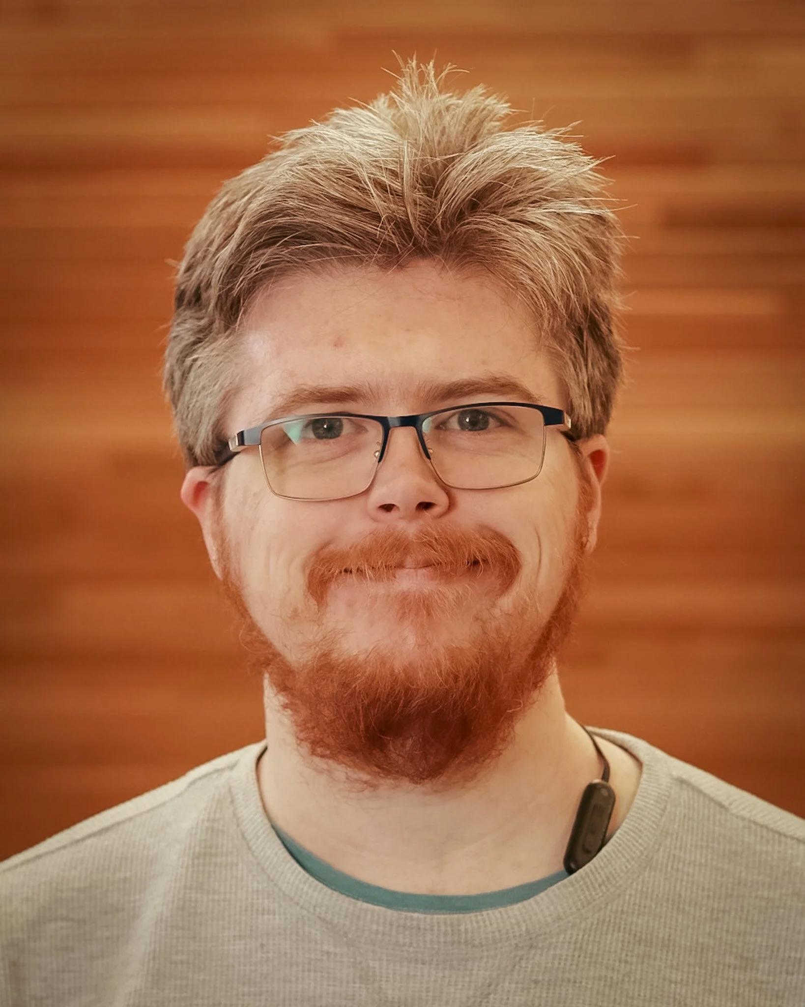 A man with glasses, red hair, and a beard smiling in front of a wooden background.