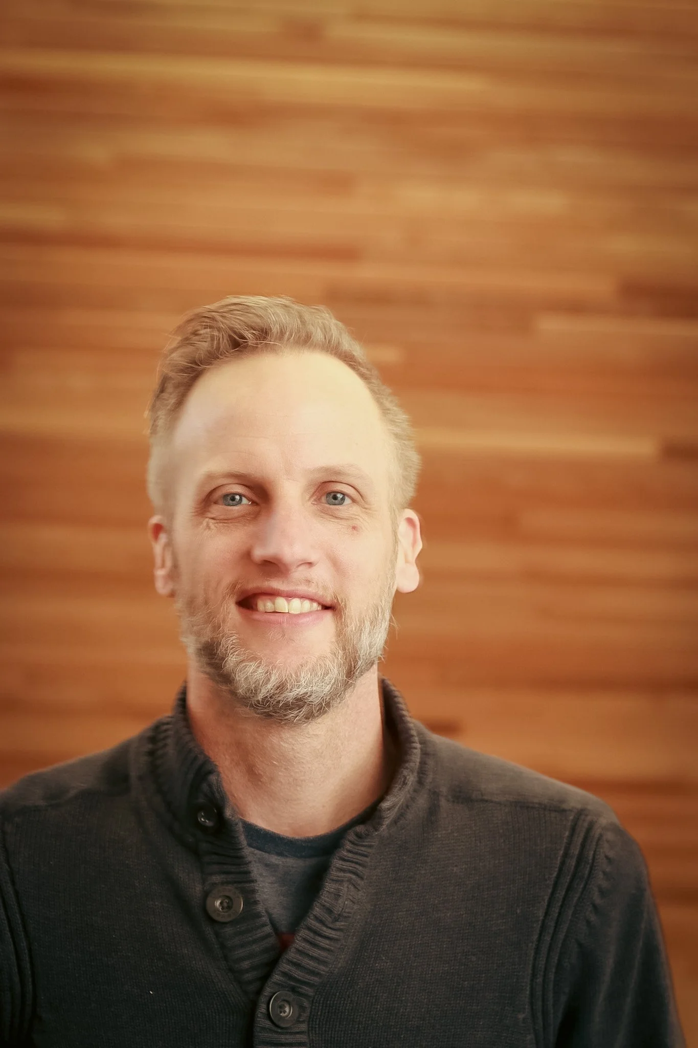 A man with blue eyes, light facial hair, and short, light brown hair smiling against a wooden background.