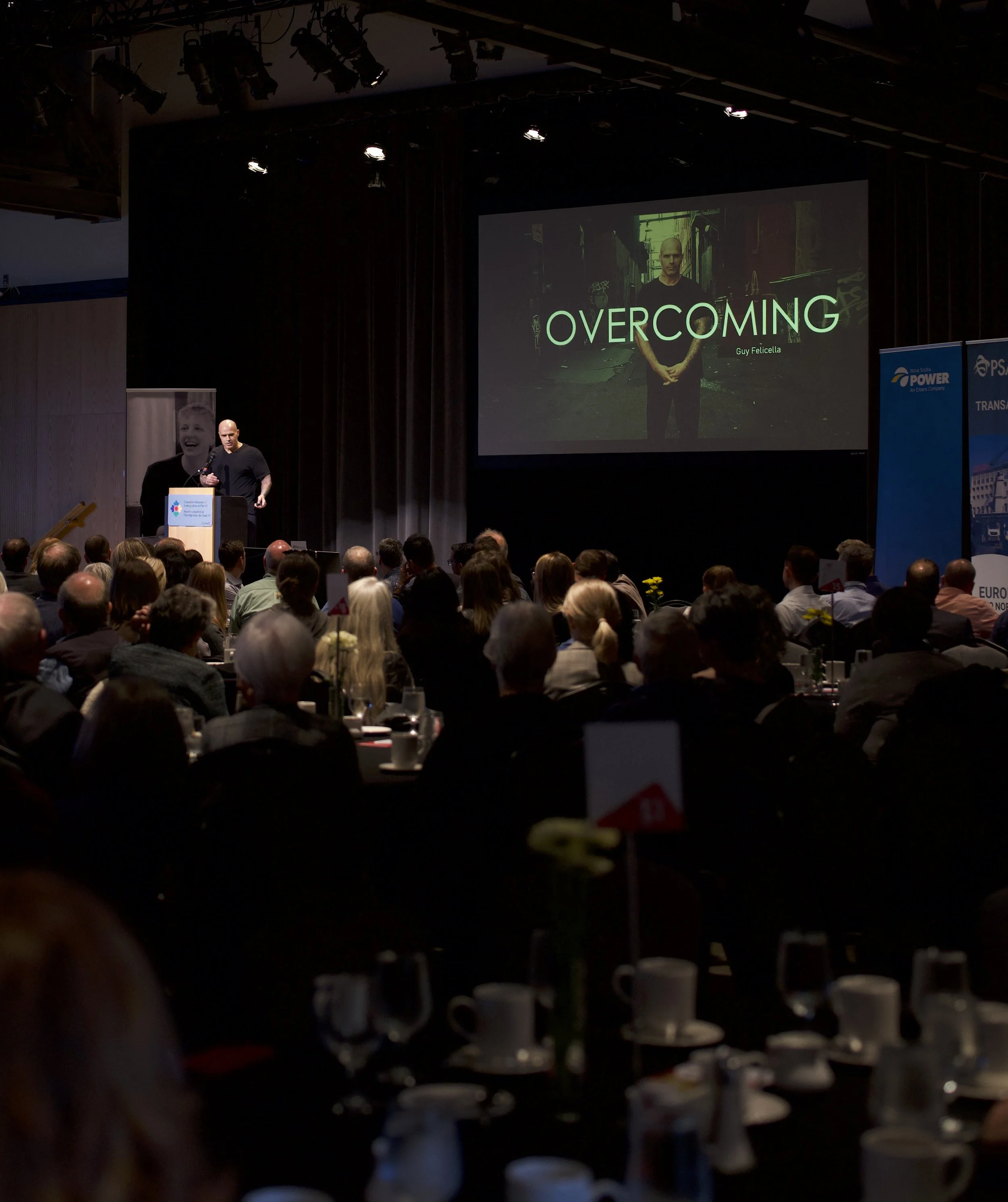 A presentation on stage titled "Overcoming" by Guy Felicella, with an audience seated in front, a large projection screen displaying his image, and stage lighting overhead in a conference setting.