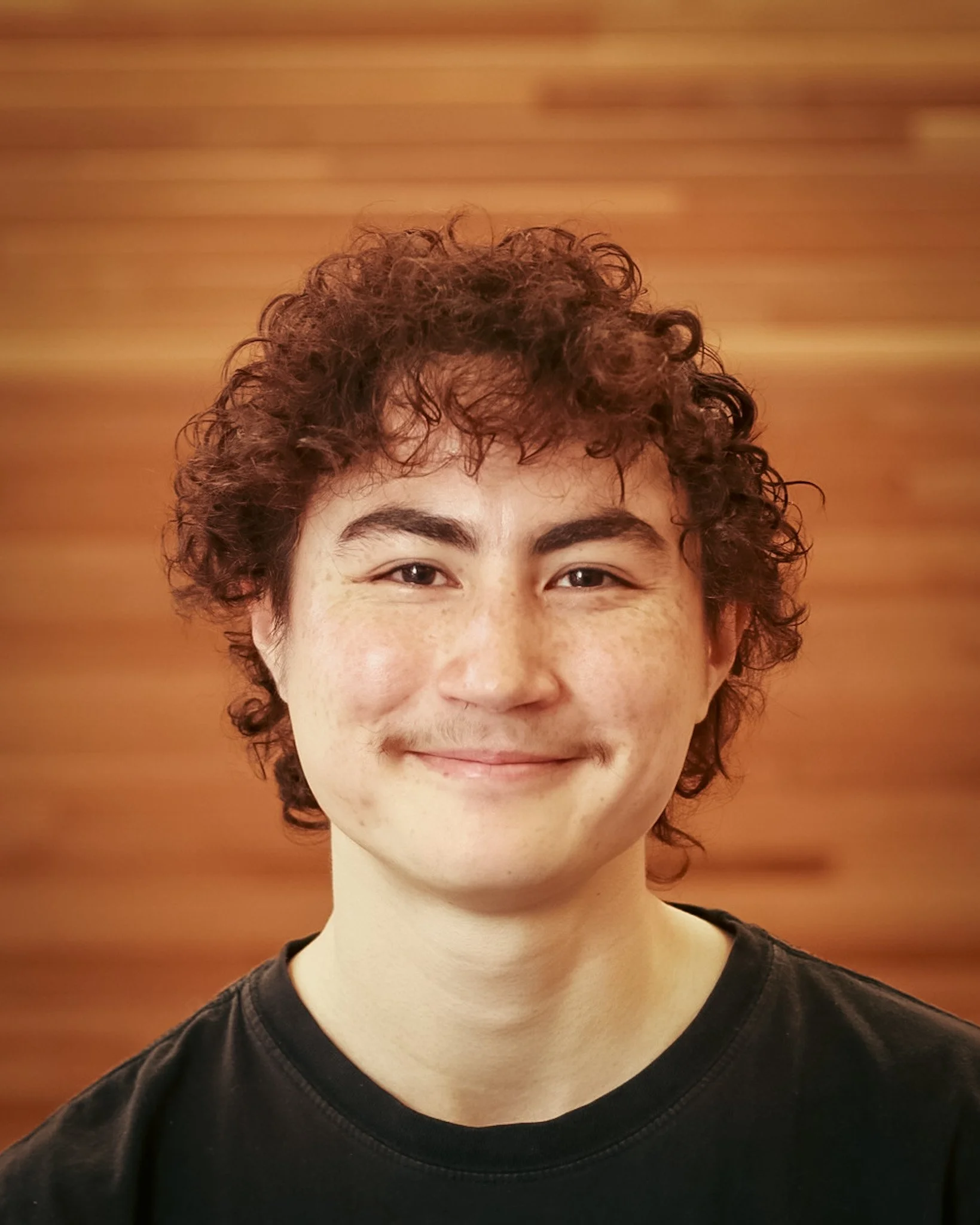 A young person with curly hair smiling against a wooden wall background.