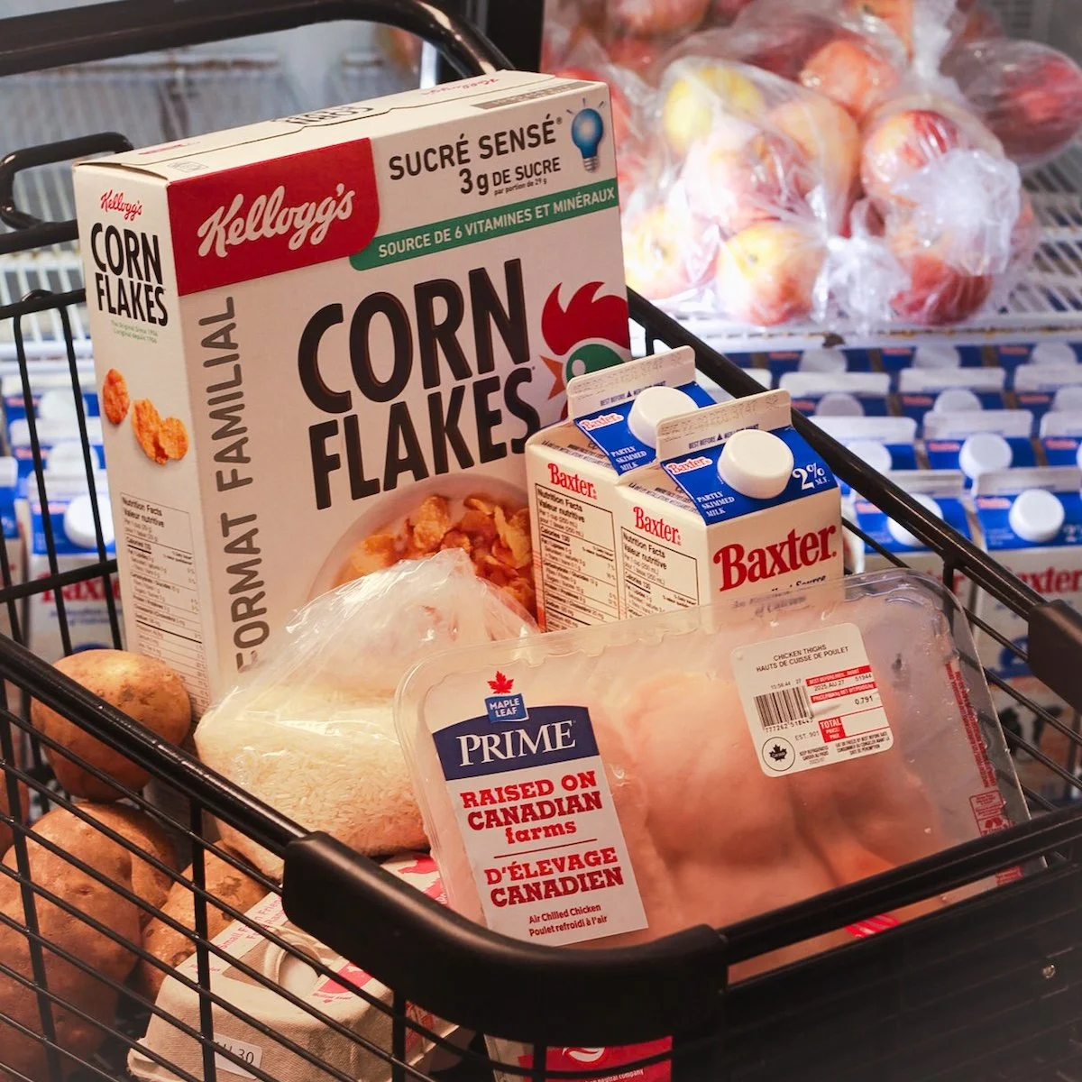 Grocery cart containing a box of Kellogg's Corn Flakes, two cartons of Baxer non-dairy creamer, a package of raw chicken, and potatoes, with a bag of apples in the background.