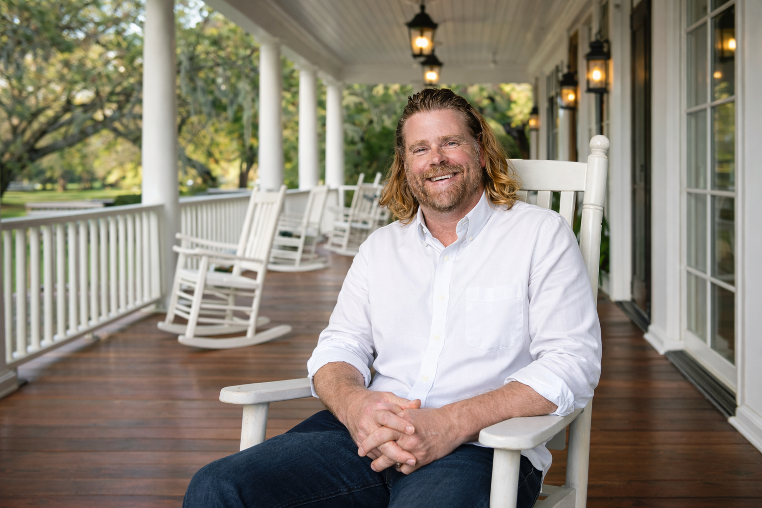 A smiling man with long hair and a beard sitting on a white rocking chair on a porch with multiple white rocking chairs behind him, surrounded by trees.