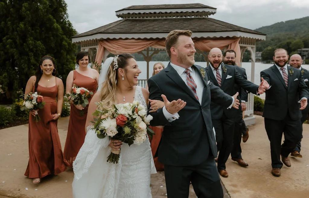 A wedding party walks outside near a gazebo with a lake and hills in the background. The bride, wearing a white dress and holding a bouquet, smiles as she walks with the groom and wedding attendants dressed in formal attire.