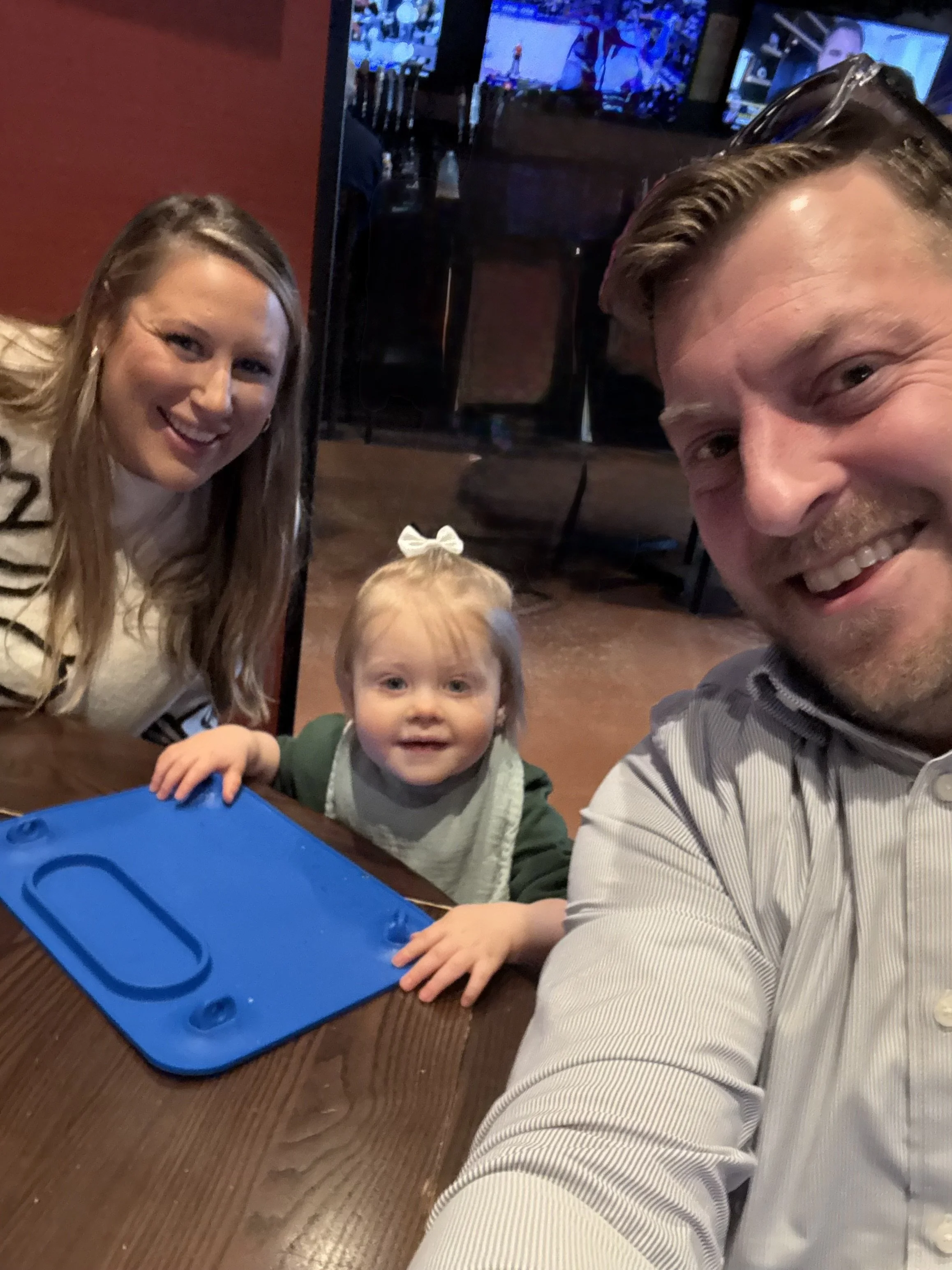 A family of three taking a selfie at a restaurant, with a woman, a man, and a young girl with a bow in her hair smiling at the camera.