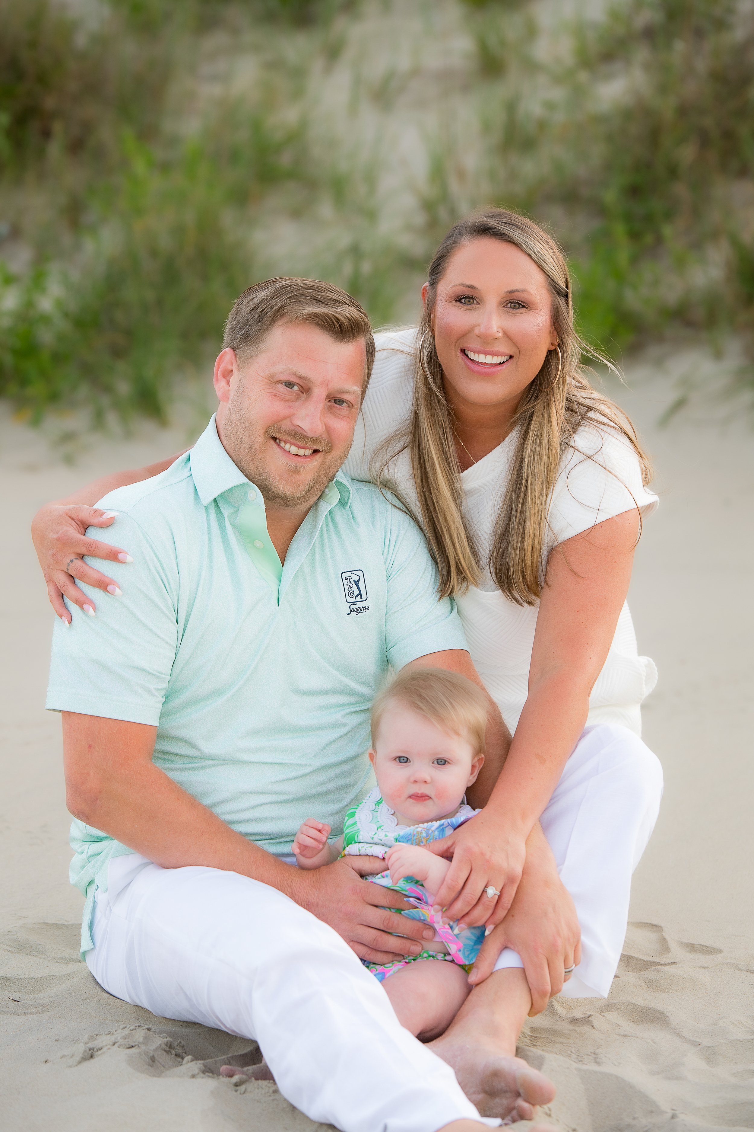 Happy family with smiling woman, man, and baby girl sitting on sandy beach with green plants in background.