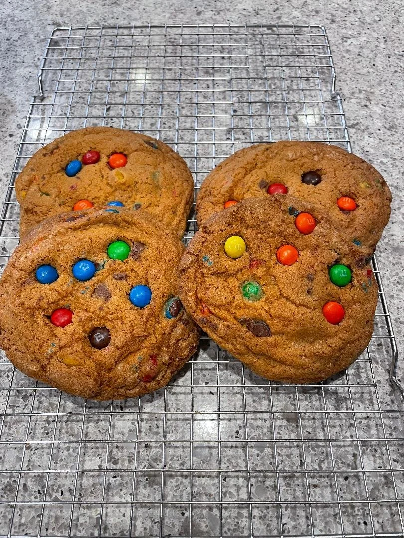 Four large chocolate chip cookies with colorful candy-coated chocolates on a wire cooling rack on a gray speckled countertop.