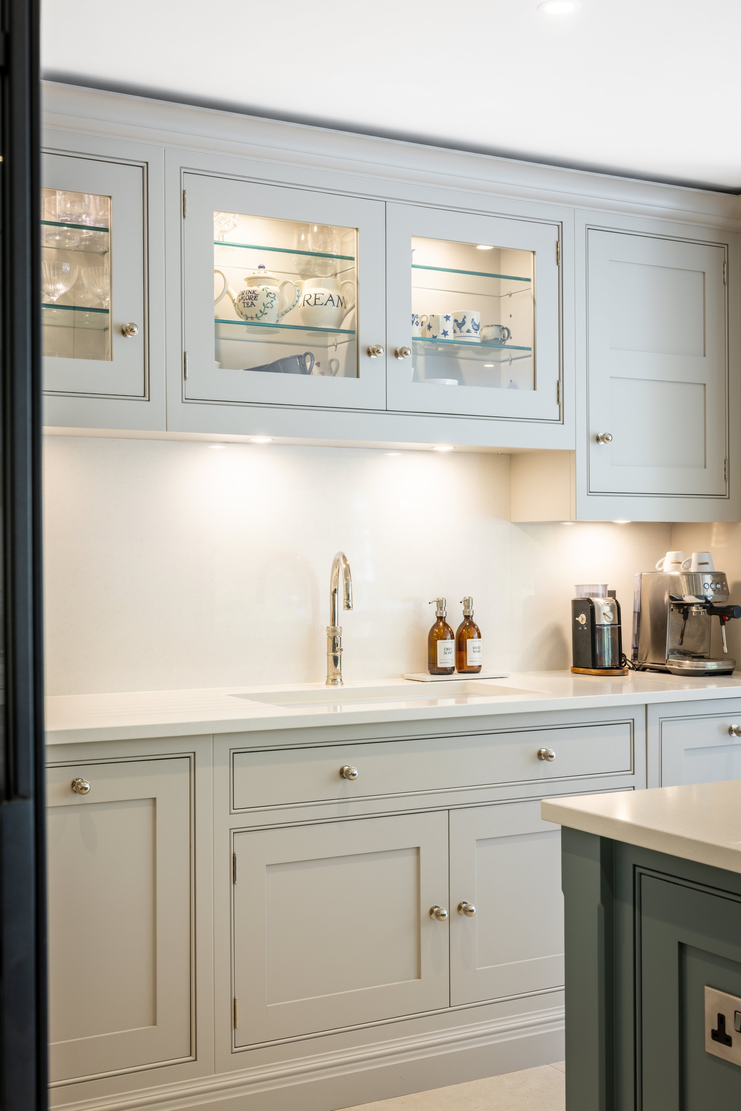 Kitchen sink and chrome tap beneath pale cabinetry and a glazed display cupboard.