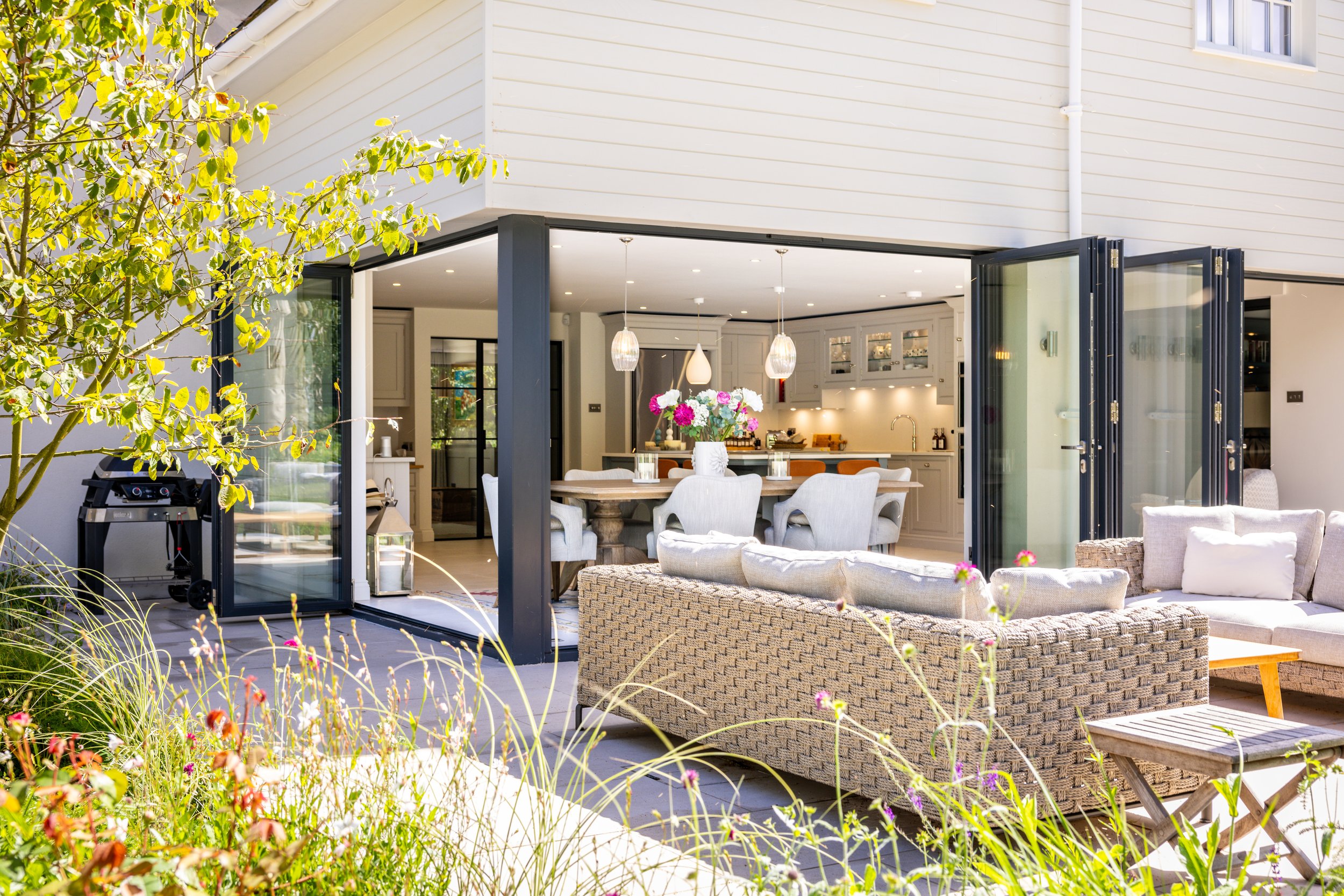 Angled view of the open kitchen doors, pale terrace paving, outdoor seating and planting.