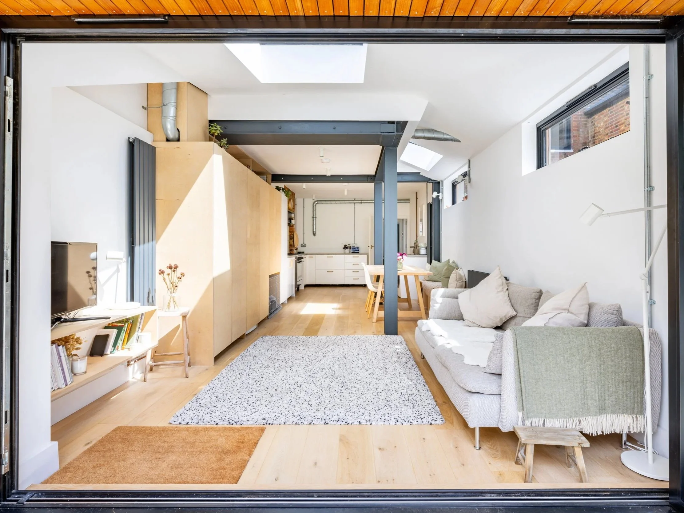 View through Town End’s open garden doors into a bright living, dining and kitchen extension with exposed dark framing and rooflights.