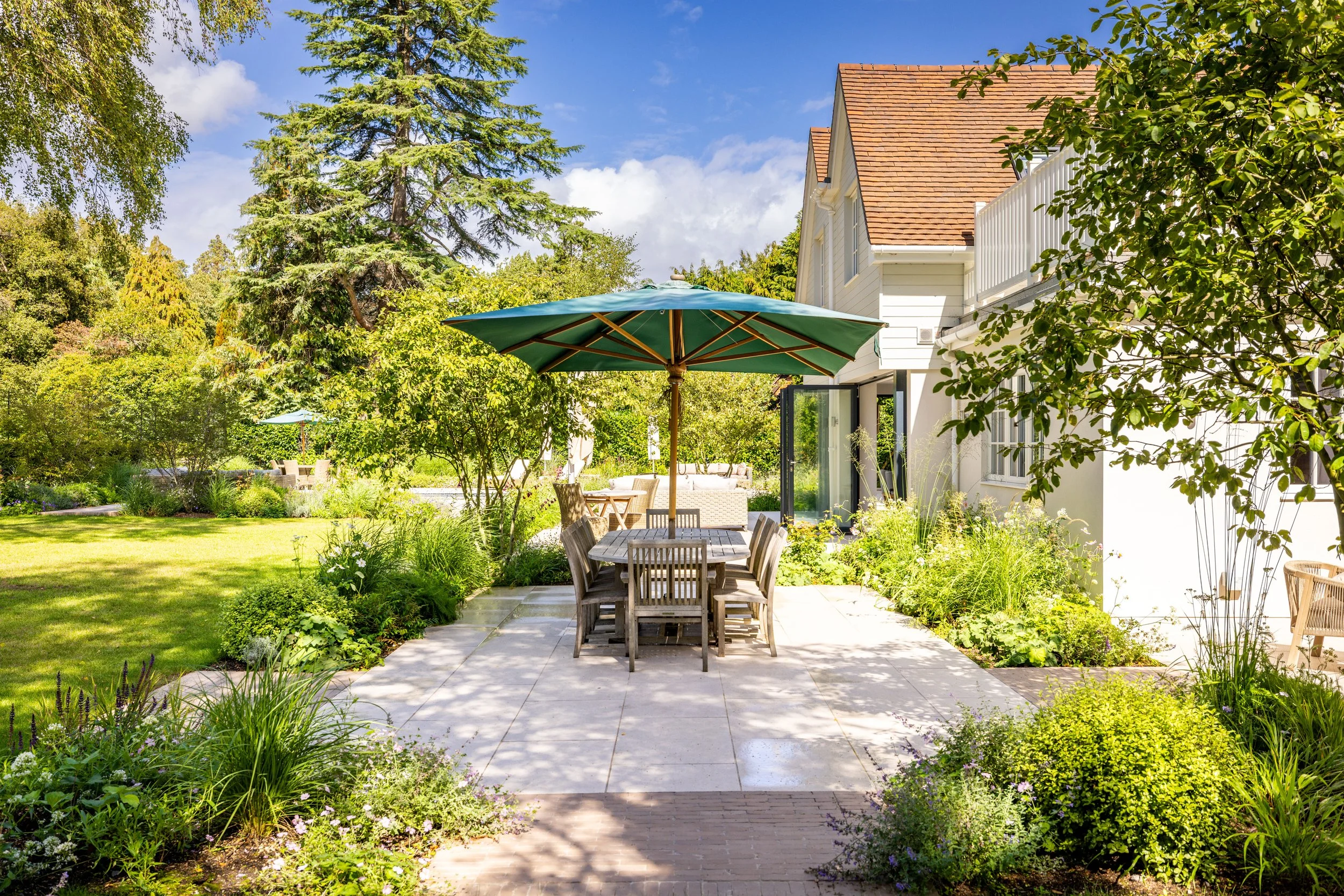 Outdoor dining terrace with a green umbrella, mature planting and the house beyond.
