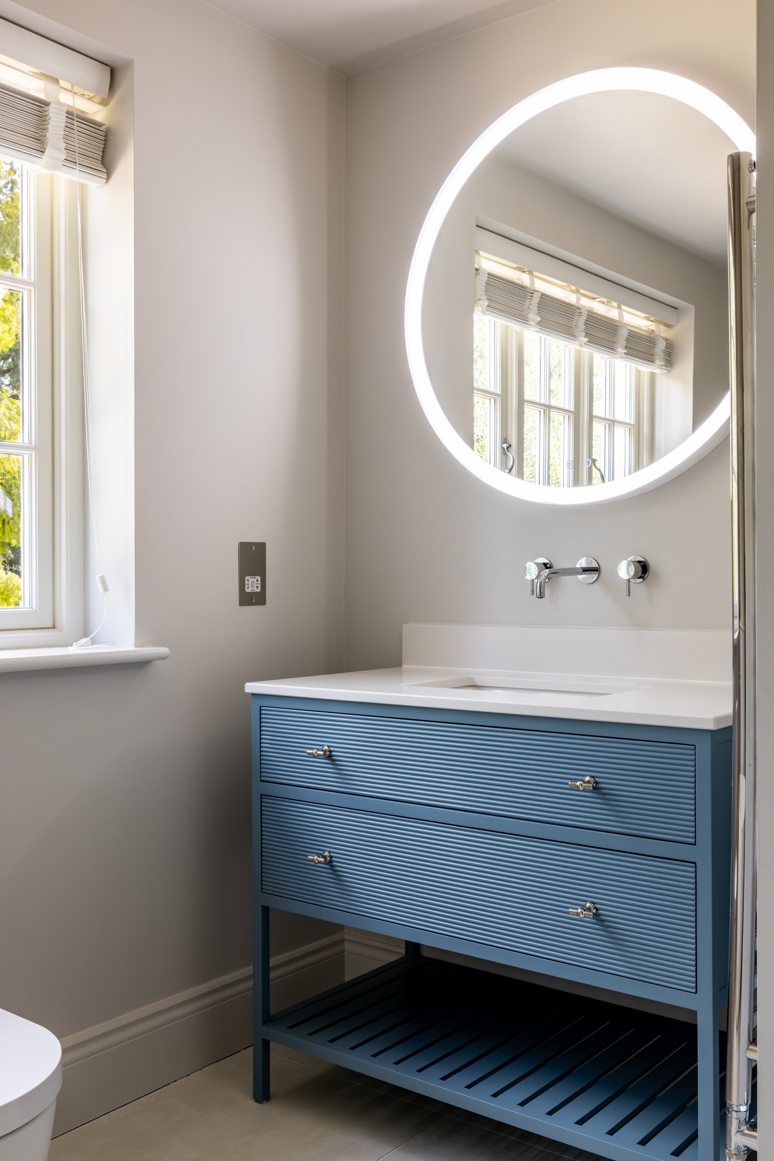 Bathroom vanity with a round vessel basin and illuminated circular mirror beside a window.