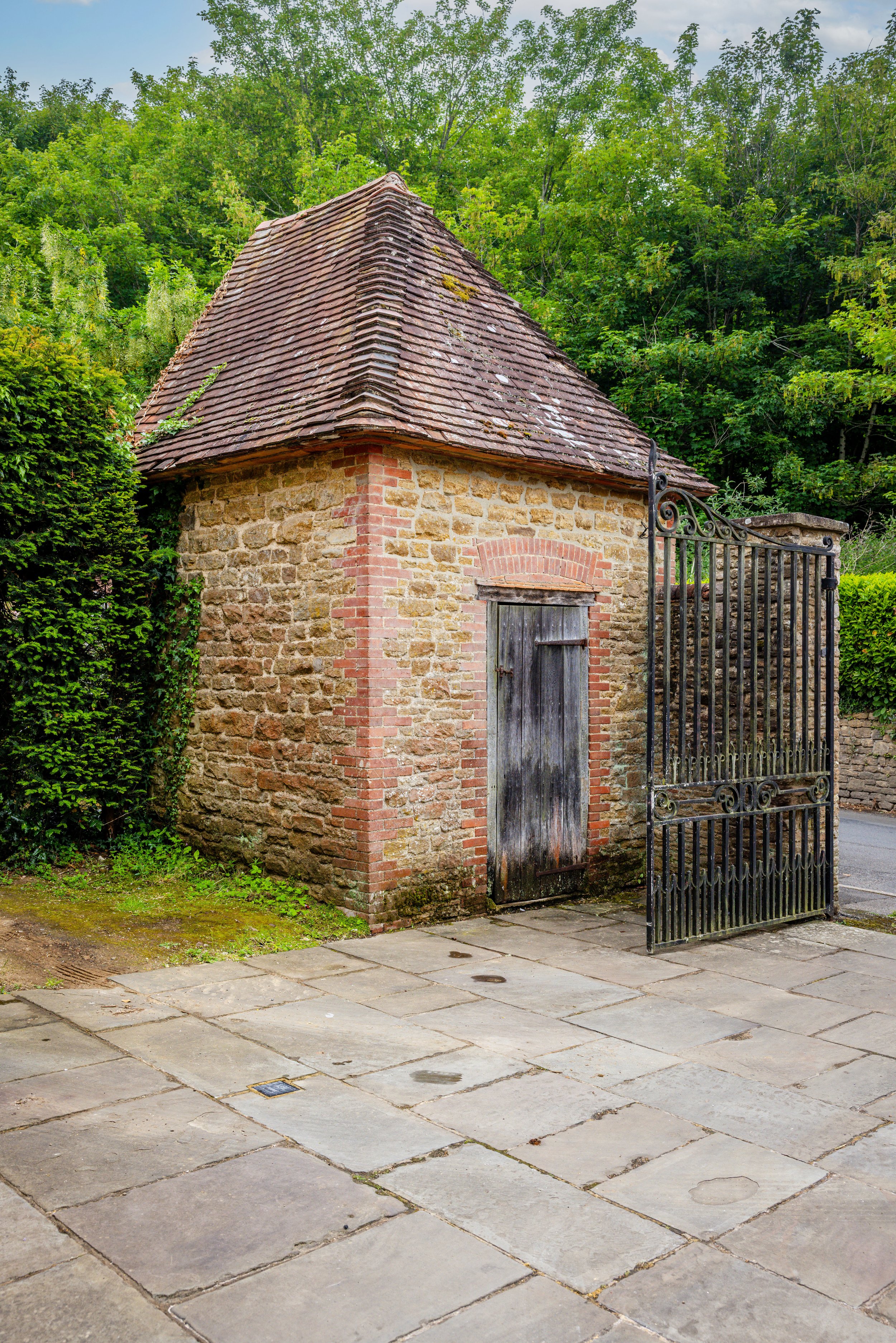 Stone garden building with a tiled roof, timber door and open metal gate.