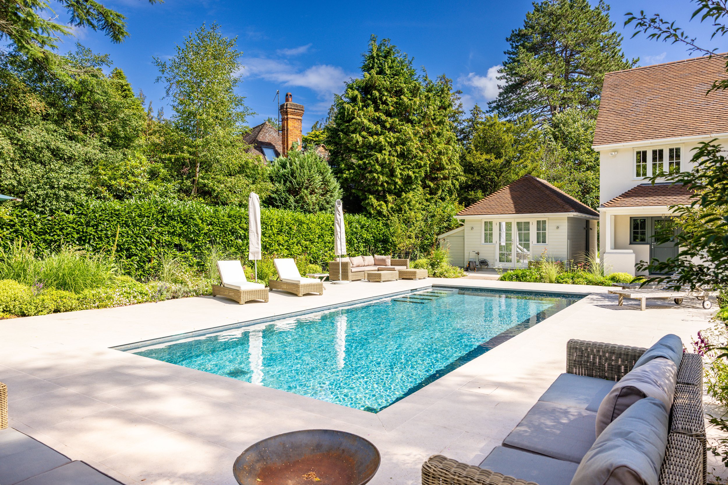View across the swimming pool towards the house, pale terrace paving and outdoor sofas.