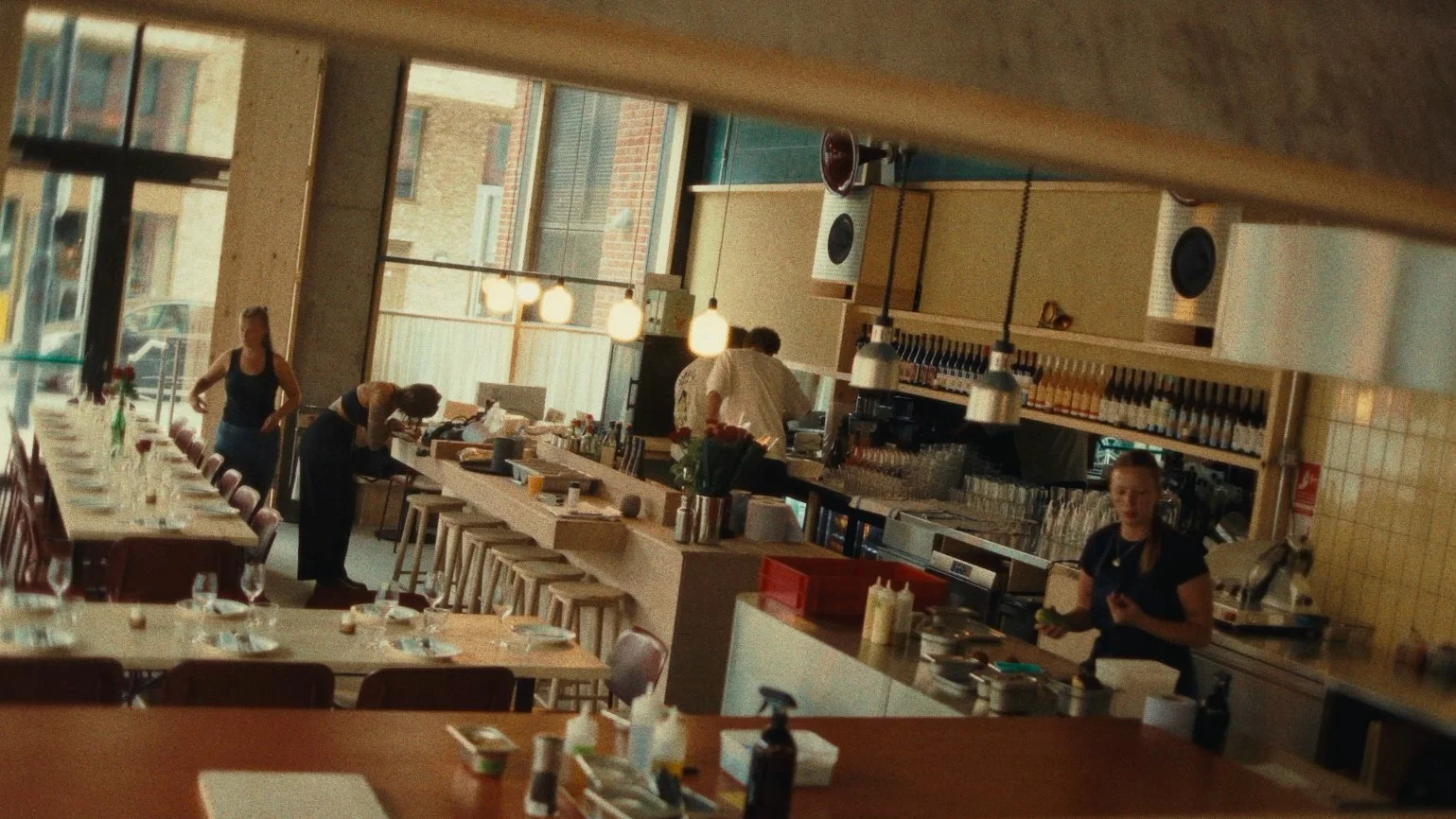 Inside a restaurant with a long bar and tables, staff preparing food, large windows letting in natural light, and modern decor.